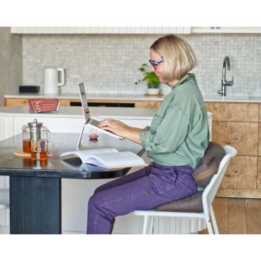 Uśmiechnięta kobieta pracująca przy stole, siedząca na brązowej poduszce klin i oparta o wałek – komfort w domowym biurze.